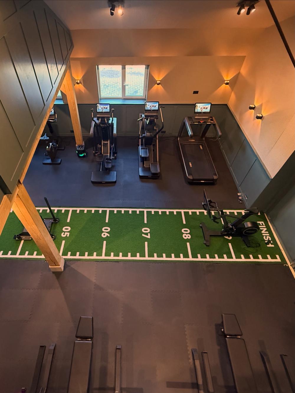 De studio van boven gezien: een groene sprintbaan met kantoorlijnen, cardioapparaten langs de wand en eikenhouten balken